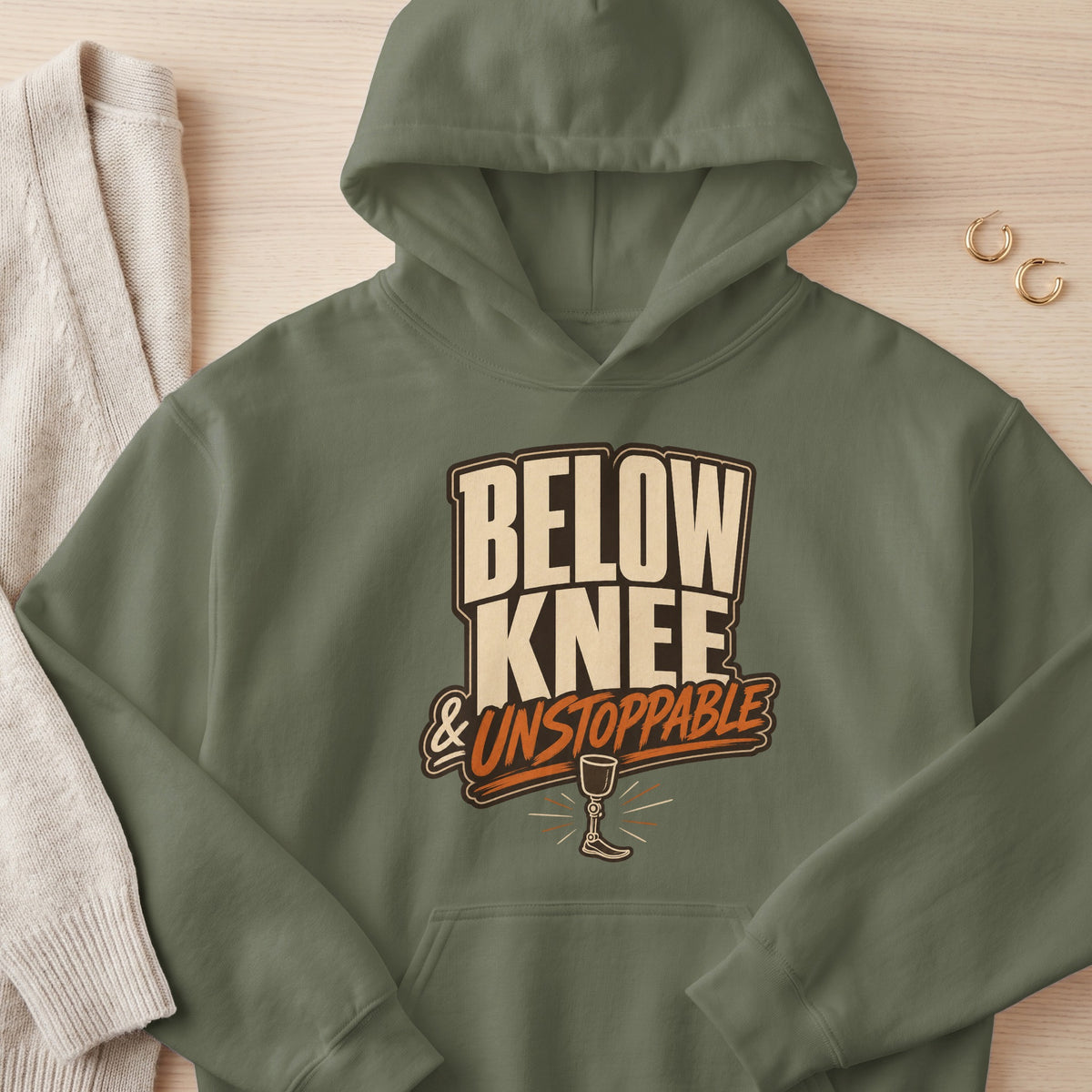 Olive green hoodie with "BELOW KNEE & UNSTOPPABLE" text and prosthetic leg graphic.