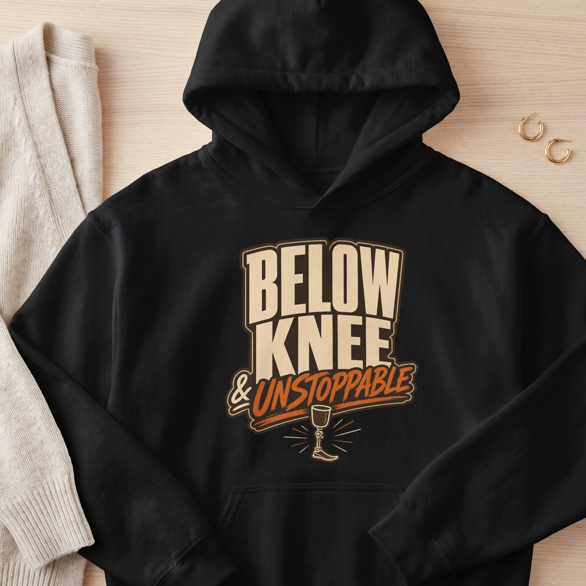 Black hoodie with "BELOW KNEE & UNSTOPPABLE" text and prosthetic leg graphic.