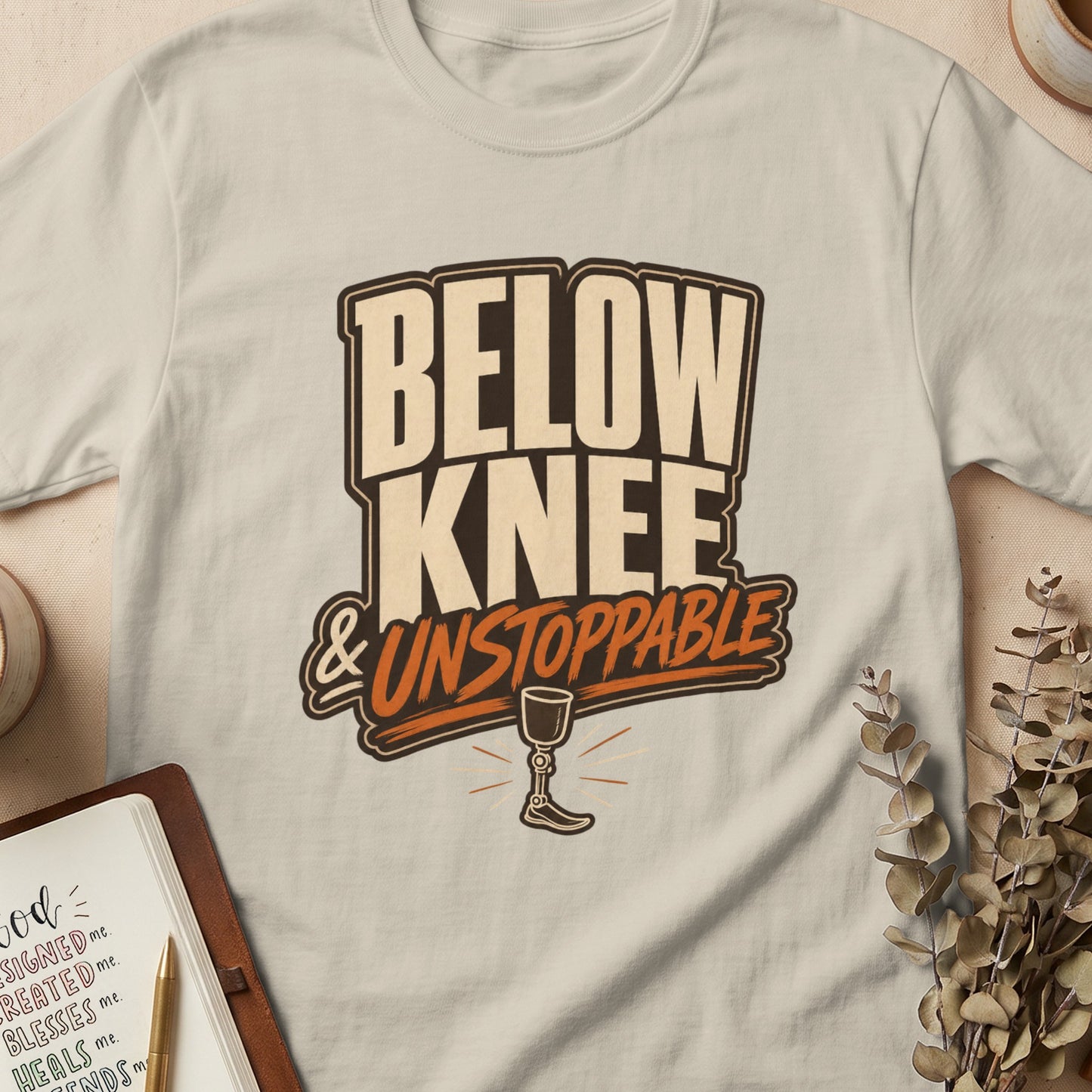 Cream t-shirt with "BELOW KNEE & UNSTOPPABLE" text and prosthetic leg graphic.