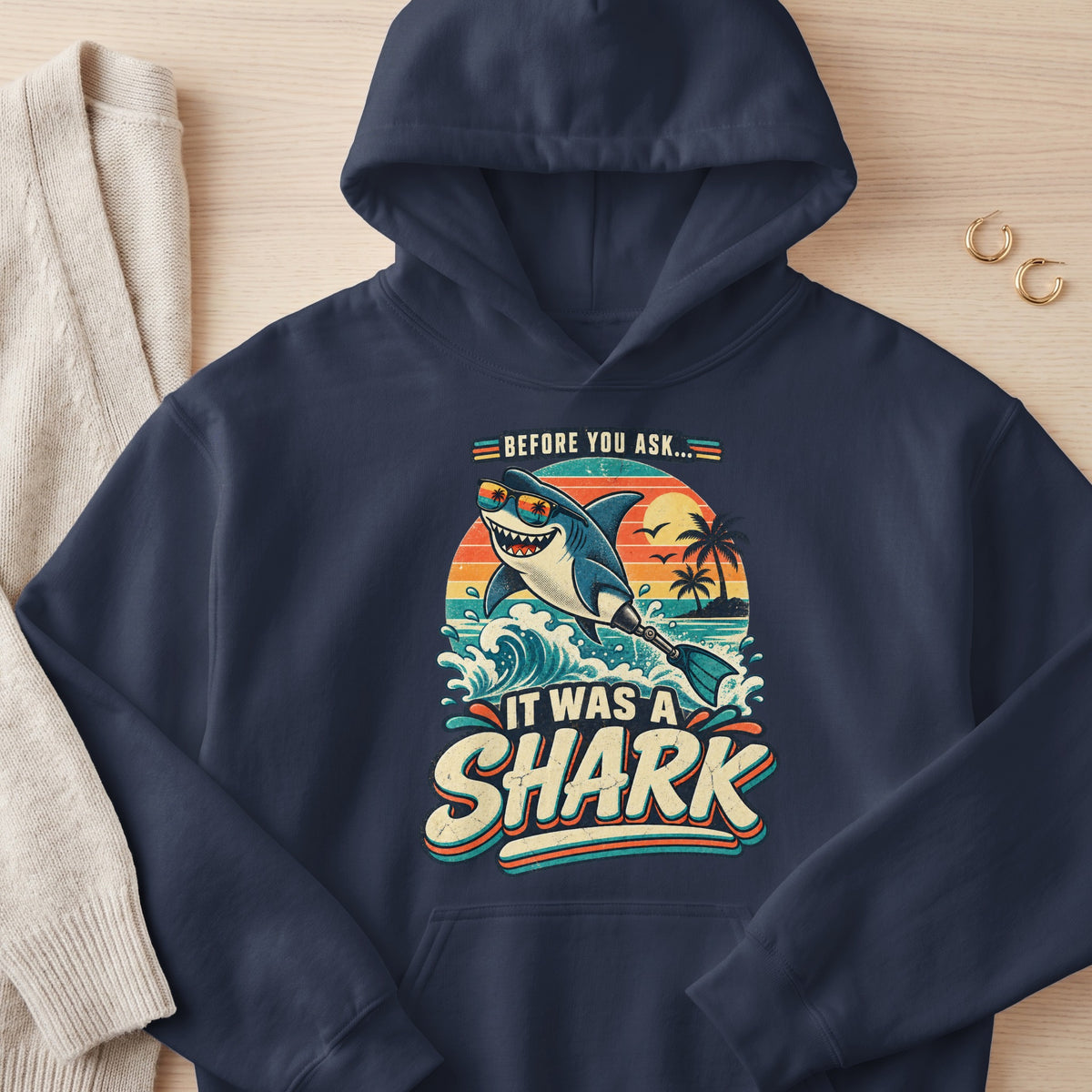 Navy hoodie with graphic of shark with sunglasses and a prosthetic tail, text reads "Before you ask... It was a shark."