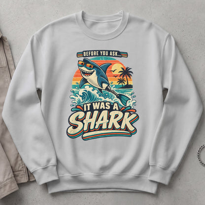 White sweatshirt with a graphic of a smiling shark wearing sunglasses and the text "IT WAS A SHARK".