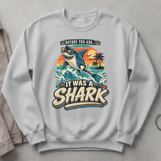 White sweatshirt with a graphic of a smiling shark wearing sunglasses and the text "IT WAS A SHARK".