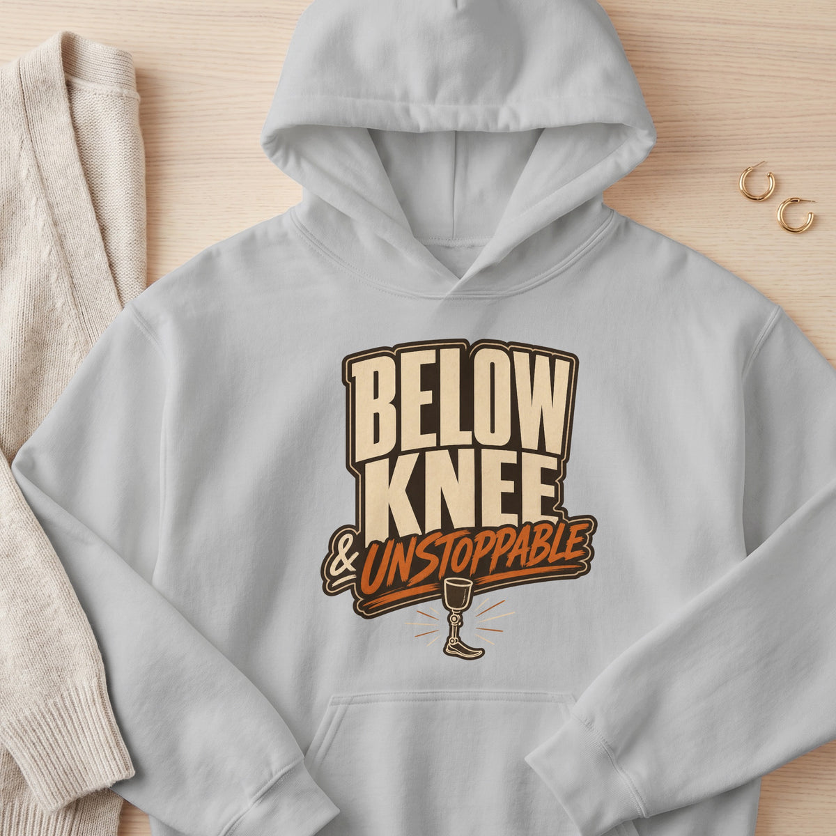 Grey hoodie with "BELOW KNEE & UNSTOPPABLE" text and prosthetic leg graphic.